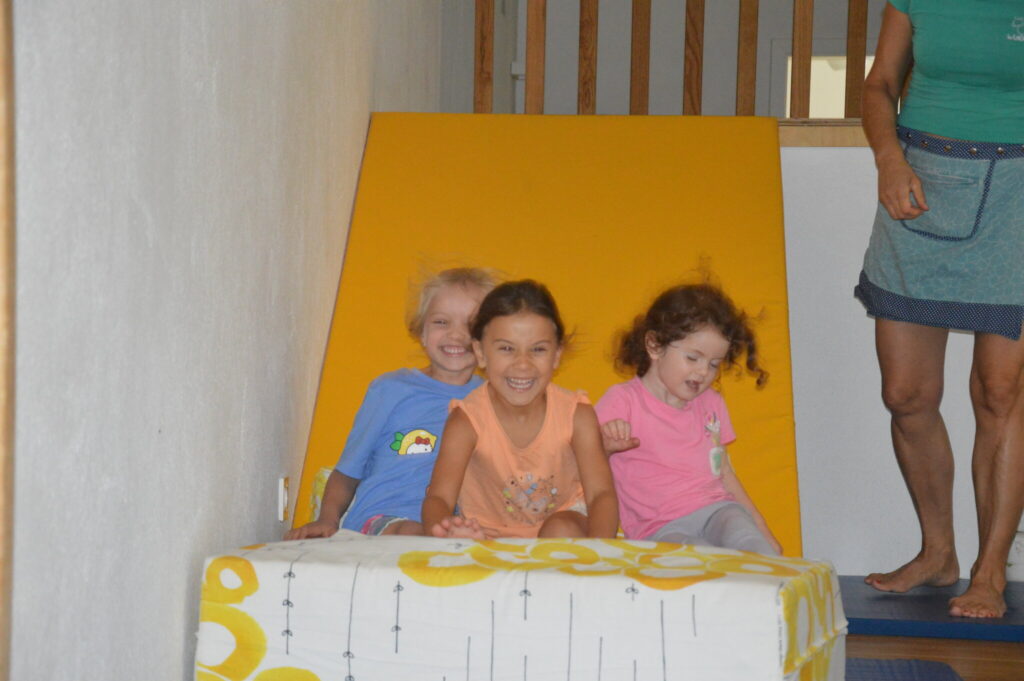Drei Kinder sitzen lachend am Fuß einer gelben Rutsche
