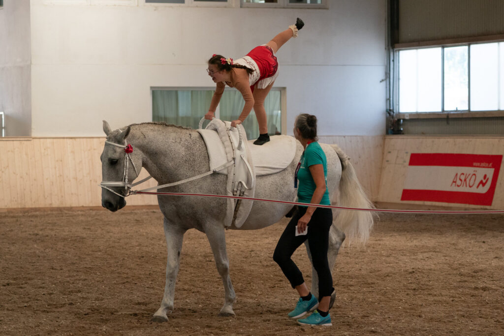Voltigiererin im Handstand auf einem galoppierenden Schimmel, Longenführerin daneben